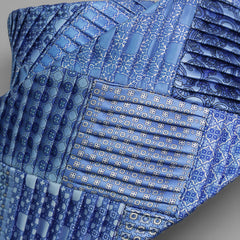ブルー系のシルク生地を組み合わせた、プリーツ仕立てのパッチワークネクタイの接写。
Close-up of a pleated silk patchwork tie in shades of blue, featuring intricate pattern blocks.